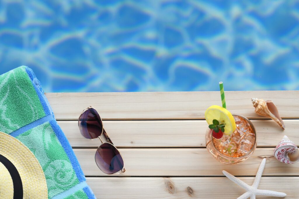 Flat lay of summer vacation travel essentials, towel sunglasses hat tropical drink by pool edge.