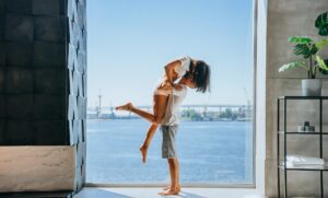 Happy newlyweds at hotel at window with view on ocean kissing. Young man lifts woman up, vacation
